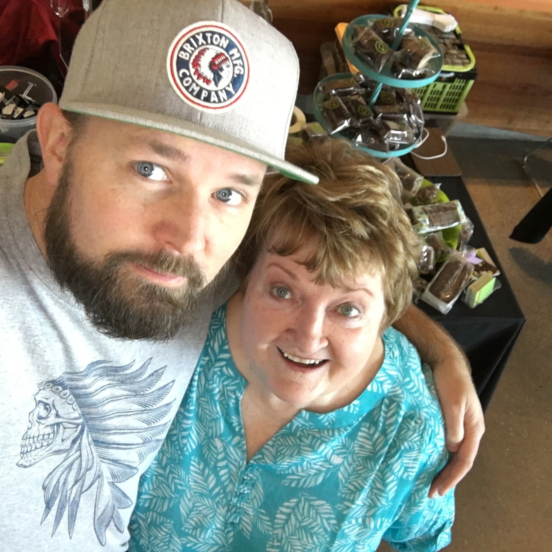 Two people posing for a selfie indoors with a casual setting. Chocolates in the background.
